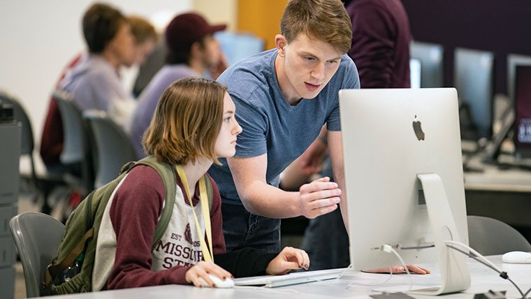 A standing Missouri State student discussing something on a Mac to a seated student. 